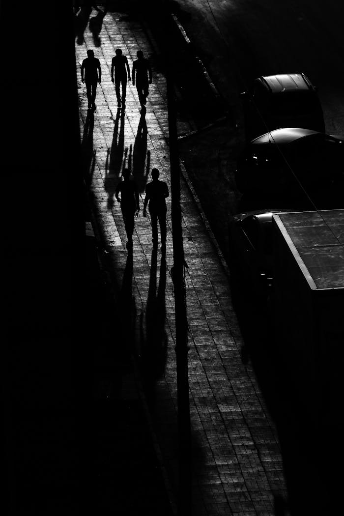Black and white scene of people casting shadows on city sidewalk at night.